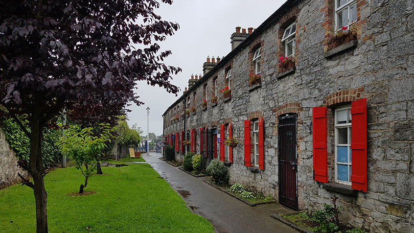 Row of two storey stone building with red shutters on right. Left, grass and trees